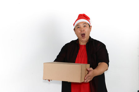 Portrait of cheerful Asian man in Santa hat holding cellphone and pointing to blank screen on white...