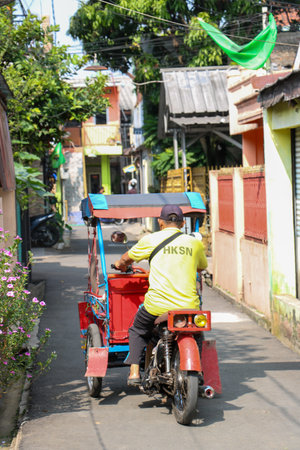 Motorized rickshaw driver carrying a heavy load of black plastic packages in an alley. Bandung,...