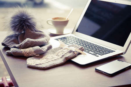 Woolen hat with pompon and mitten near a laptop and cup of coffe on the wooden tableの写真素材