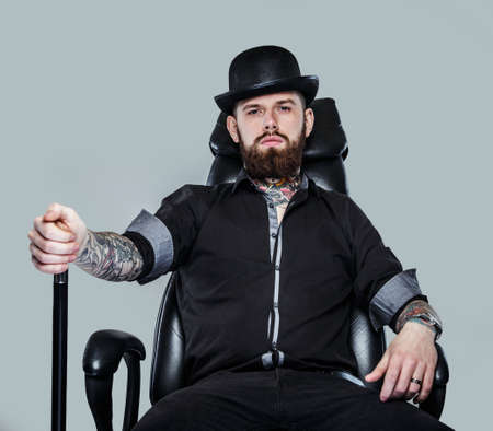Serious gangster in hat sitting on leather chair holding caneの写真素材