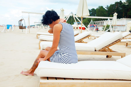 Middle age woman with black hair in striped dress sitting on a beach sofa and heaving backache.の写真素材