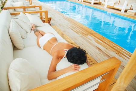 Middle age woman with black hair, tattoes on her legs  in white bikini relaxing on a beach sofa near...