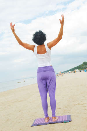 Middle age woman with shirt black hair in a white t-shirt and purple pants doing yoga on the summer beach.の写真素材