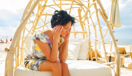 Middle age woman with shirt black hair in color dress sitting on beach sofa with headache.の写真素材