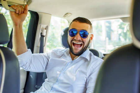 Happy man with black hair and beard, sunglasses, dressed in white shirt and blue shorts sitting on back seat of a car.の写真素材
