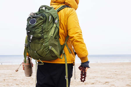Man dressed in black pants and yellow jacket from behind with green backpack stands on winter beach.の写真素材