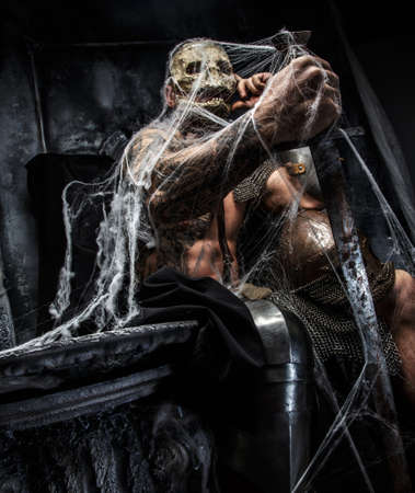 Muscular man with skull on his face and sword in his hand posing on the throne in spiders web on gray background.の写真素材