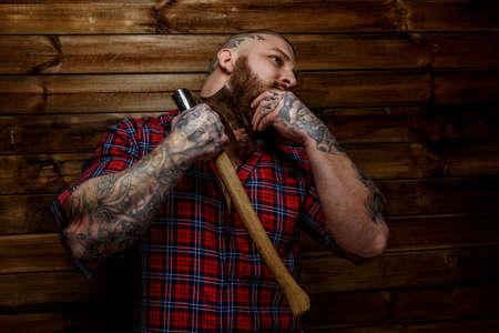 Tattoed man in red shirt in a section with beard hang axe on wooden backgroud. One hand near man face.の写真素材