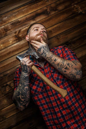 Tattoed man in red shirt in a section with beard hang axe on wooden backgroud. One hand near man face.の写真素材