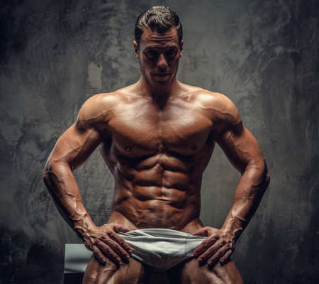 Awesome shirtles muscular guy sitting on white podium. Isolated on grey background.