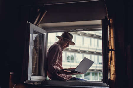 Guy in big hat sitting in the window and working with laptop.の写真素材