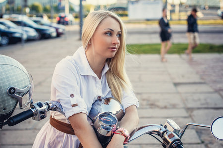 Cute girl with long blond hair in white dress possing with scooter at a city streetの写真素材