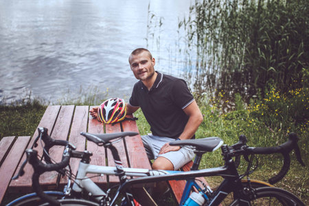 Smiling man sitting at table on lake shore with sport bycicles on background.