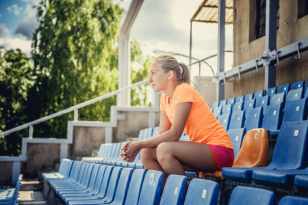 Slim female in sportswear sitting on plastic blue seat on the stadium.