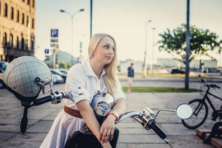 Female with long blond hair in white dress posing near scooter on street in the city.の写真素材