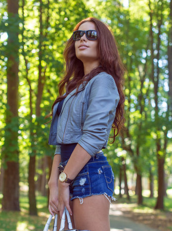 Sexy brunette woman in jeans shorts posing outside in green park.