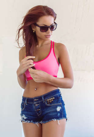 Sexy brunette female in pink top and blue jeans shorts posing over white background.の写真素材