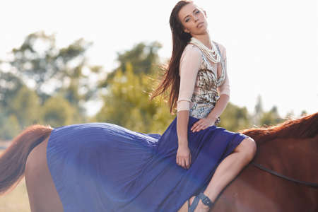 Fashion girl with long brown hair and in blue scirf sitting on brown horse's back.の写真素材