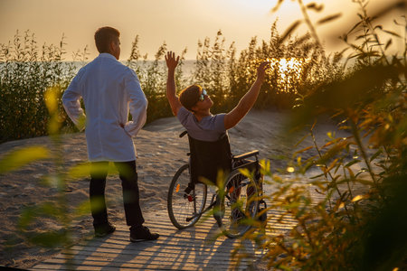 Young man in wheelchair and his doctor relaxing on the beach over sunset.の写真素材