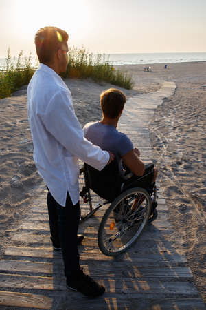 Young man in wheelchair and his doctor relaxing on the road to the beach.の写真素材