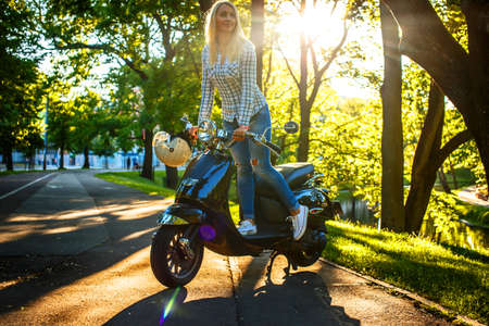 Awesome blond female in jeans and shirt posing on scooter in green park.の写真素材