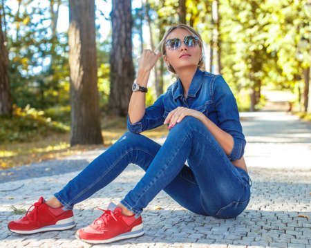 Slim blond female in jeans costume and red shoes sitting on the road in a park.の写真素材