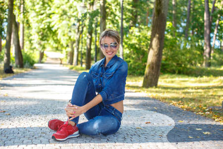 Beautiful girl in jeans pants and jeans jacket sitting on the road in a park.の写真素材