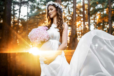 Pregnant woman in long white dress holding bouquet from pink flowers. Sunset behind tree in the forest.の写真素材