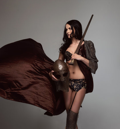 brunette woman in ancient armor holding helmet and sword. Isolated on grey background.の写真素材