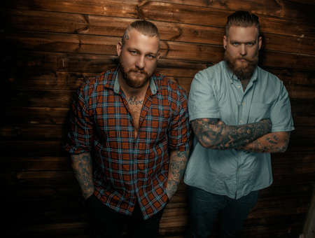 Two serious mans with beards and tattoos posing over wooden wall.