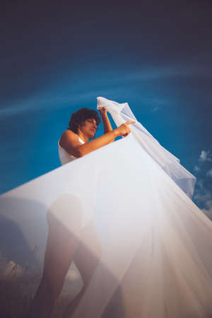 Brunette middle age woman in white underwear holding fluttering cloth over blue sky with white clouds.の写真素材