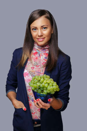 Smiling caucasian woman in a suit holds grape. Isolated on grey background.の写真素材