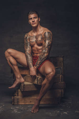 Naked tattooed athletic man sitting on wooden boxes in studio.の写真素材