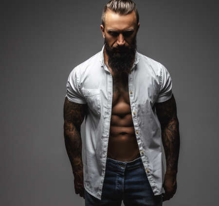 Serious bearded man with tattooes on his arms in a white shirt. Isolated on a grey background.の写真素材