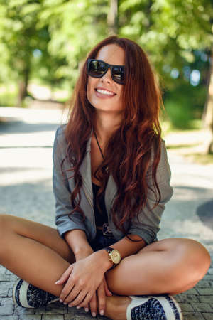 Awesome young brunette woman in a sunglasses sitting on a park road.の写真素材