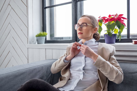 A woman in eyeglasses seats near the window at home.