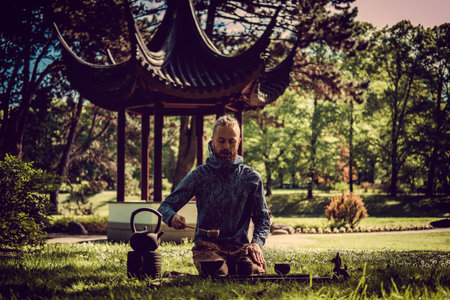 Caucasian man performed chinese tea ritual in summer park with traditional chinese arbour on background.の写真素材