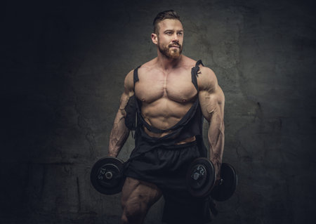 Powerful huge bodybuilder with dumbbells posing on dark grey background in a studio.の写真素材