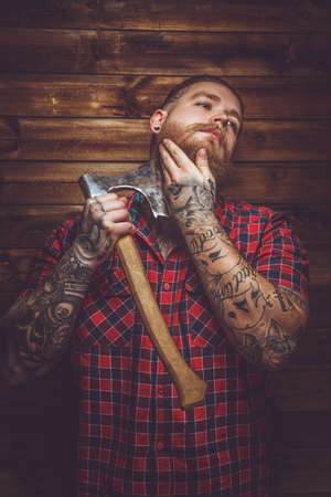 Tatooed  bearded maleholding axe and posing over wooden wall in a studio.の写真素材