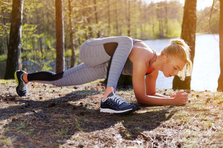 Sporty blond female doing fitness exercises near wild river in warm summer day.