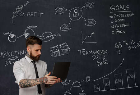 Tattooed bearded techer with laptop over school desk with formulas.