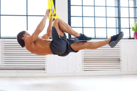 Sporty male exercising with fitness trx straps over big bright window background.