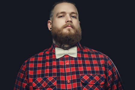 Tattooed bearded unformal male in red shirt and grey bow tie isolated on a darg grey background.の写真素材