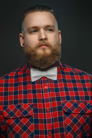 Tattooed bearded unformal male in red shirt and grey bow tie isolated on a darg grey background.