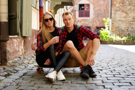 Blond attractive female in sunglasses and her boyfriend sitting on longboard on a street in old town.の写真素材