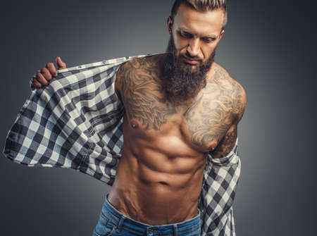 Tattooed bearded male taking off his shirt and undressing for studio shooting.の写真素材