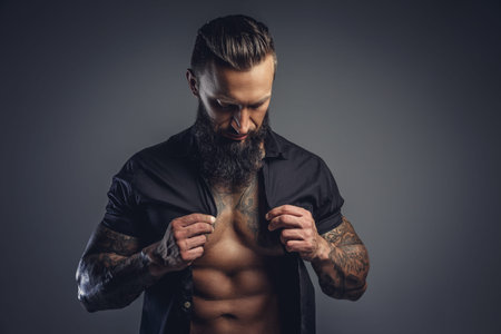 Tattooed bearded male taking off his shirt and undressing for studio shooting.