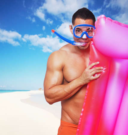 Portrait of muscular male in underwater scuba mask holding pink water matress on a beach under blue sky.の写真素材