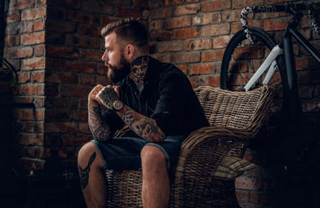 Portrait of a bearded hipster male relaxing in a chair with fix bicycle and the wall from the red brick background.の写真素材