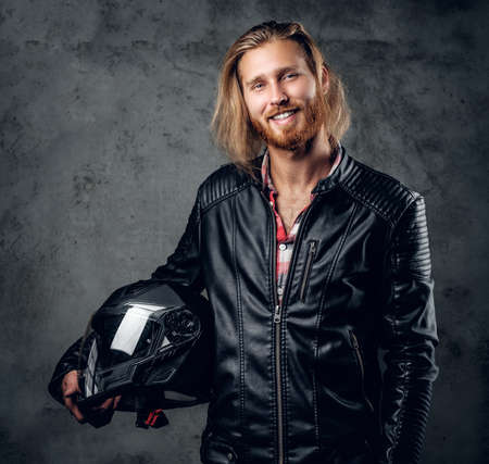 Positive bearded redhead male in leather jacket holds motorcycle helmet on grey background.の写真素材